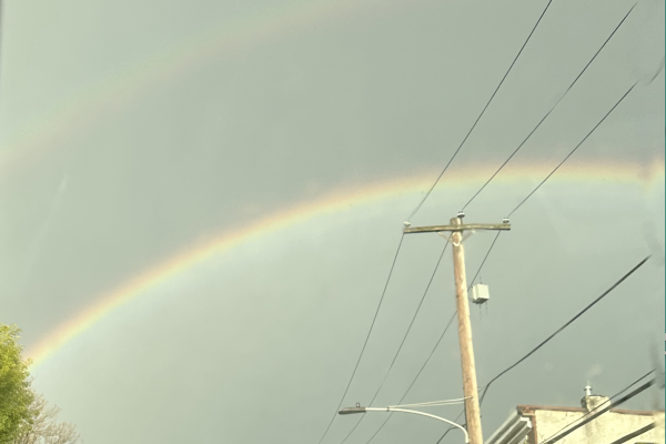 A double rainbow appears in a cloudy sky above utility poles and buildings.